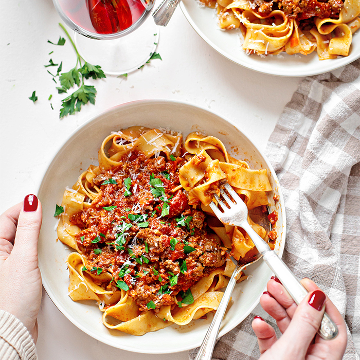 Overhead shot of a woman enjoying a bowl of pasta Bolognese with a fork on Valentine's Day, paired with a glass of red wine. Two bowls of hearty pasta sit on the table, creating a cozy and romantic dinner setting. Perfect for an easy Valentine’s Day dinner idea