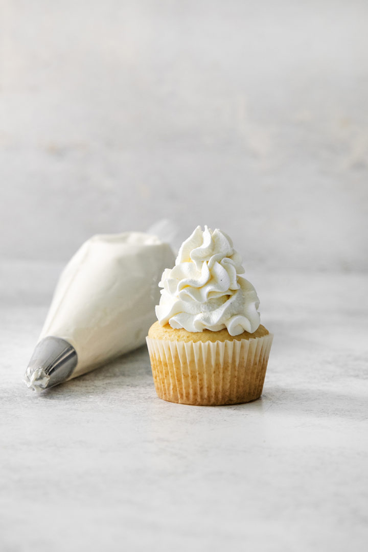 A vanilla cupcake topped with a tall swirl of stabilized whipped cream, sitting beside a piping bag with a metal tip, against a light neutral background.
