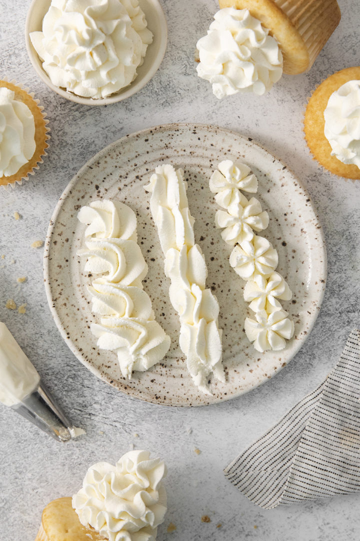 A speckled ceramic plate displaying three piped designs of whipped cream, with frosted cupcakes and a piping bag scattered around the edges.