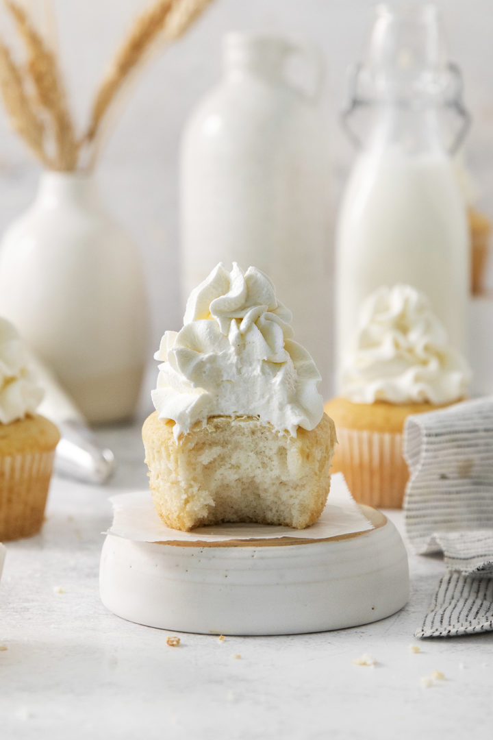 A vanilla cupcake with whipped cream frosting, shown with a bite taken out to reveal the fluffy whipped texture inside.