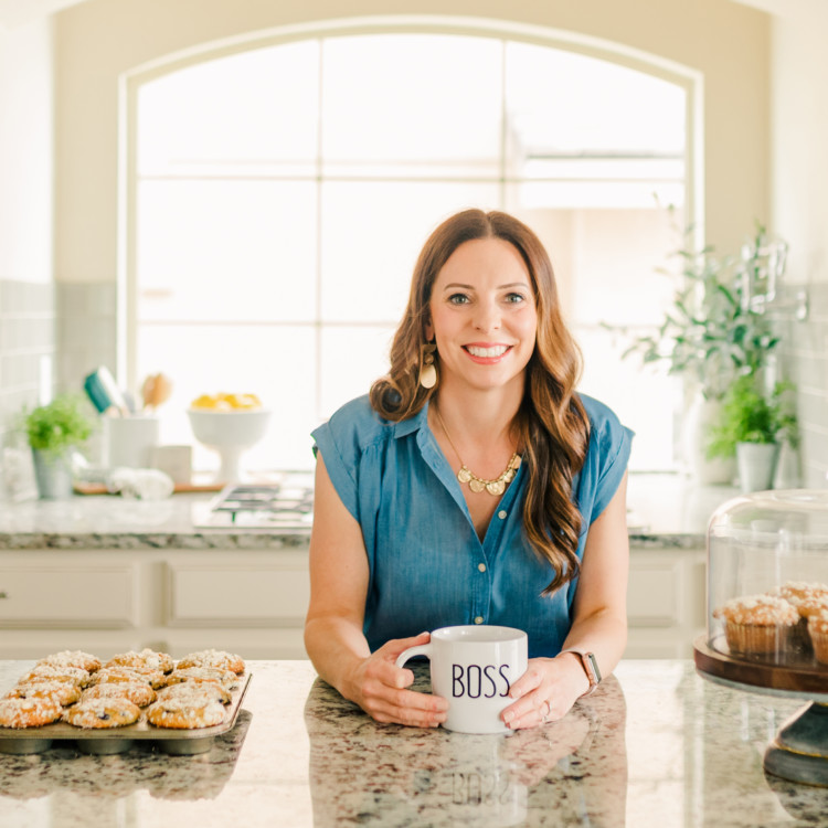 Portrait of Katie Kick leaning over a kitchen counter while holding a coffee mug labeled boss.