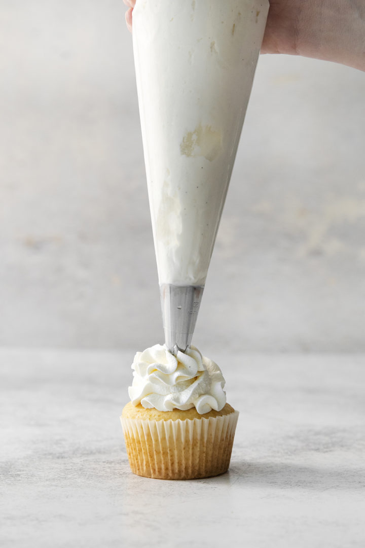 Close-up of a piping bag held vertically over a cupcake, as whipped cream is piped in a tall swirl using a star tip.