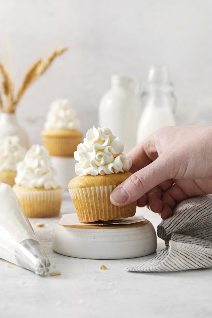 A hand gently lifts a frosted cupcake from a ceramic pedestal, with a piping bag and several more whipped cream-topped cupcakes in the background.