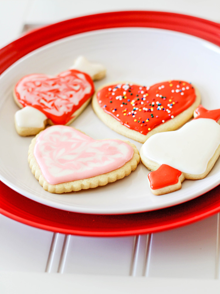 Valentine's Day cut out cookies on a red and white plate, sitting on a white wooden table. The cookies are frosted in festive red, pink, and white icing and decorated with colorful sprinkles