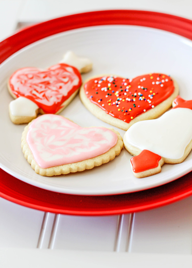 Valentine's Day cut out cookies on a red and white plate, sitting on a white wooden table. The cookies are frosted in festive red, pink, and white icing and decorated with colorful sprinkles
