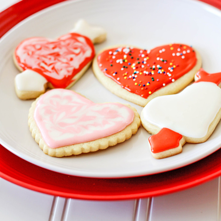 Valentine's Day cut out cookies on a red and white plate, sitting on a white wooden table. The cookies are frosted in festive red, pink, and white icing and decorated with colorful sprinkles
