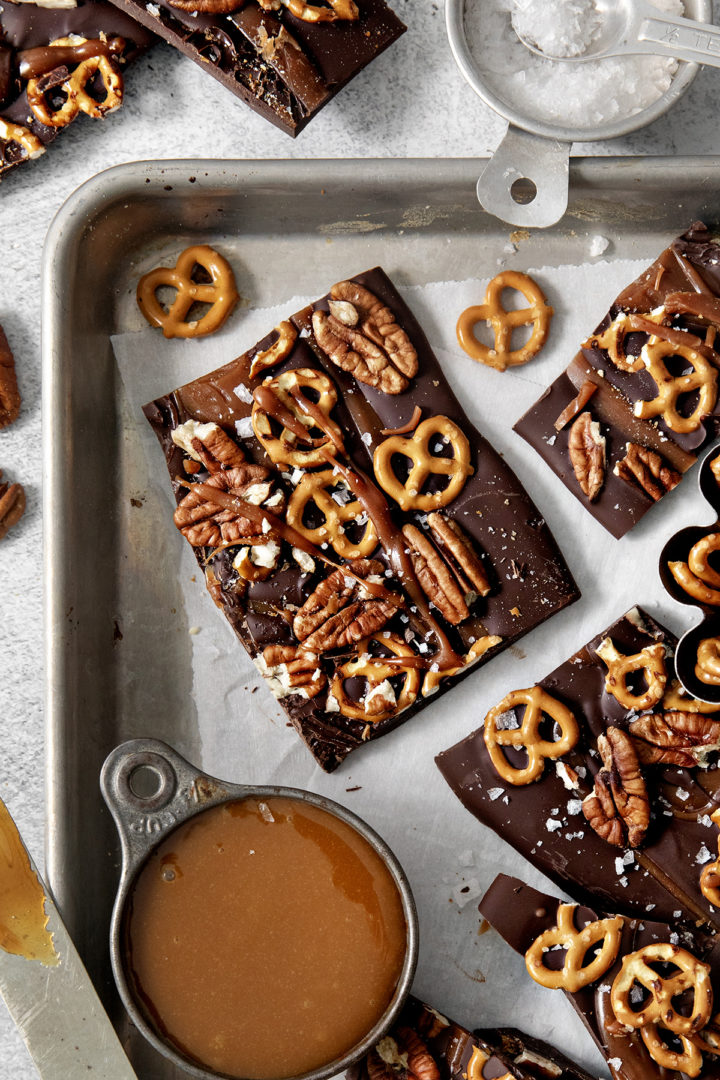 close of of a piece of dark chocolate and caramel pretzel bark next to a bowl of salted caramel