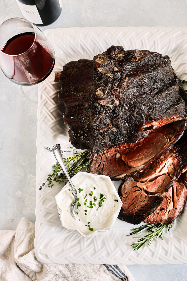 overhead photo of a serving platter with slices of smoked prime rib, prime rib sauce, and fresh herbs next to a bottle of wine