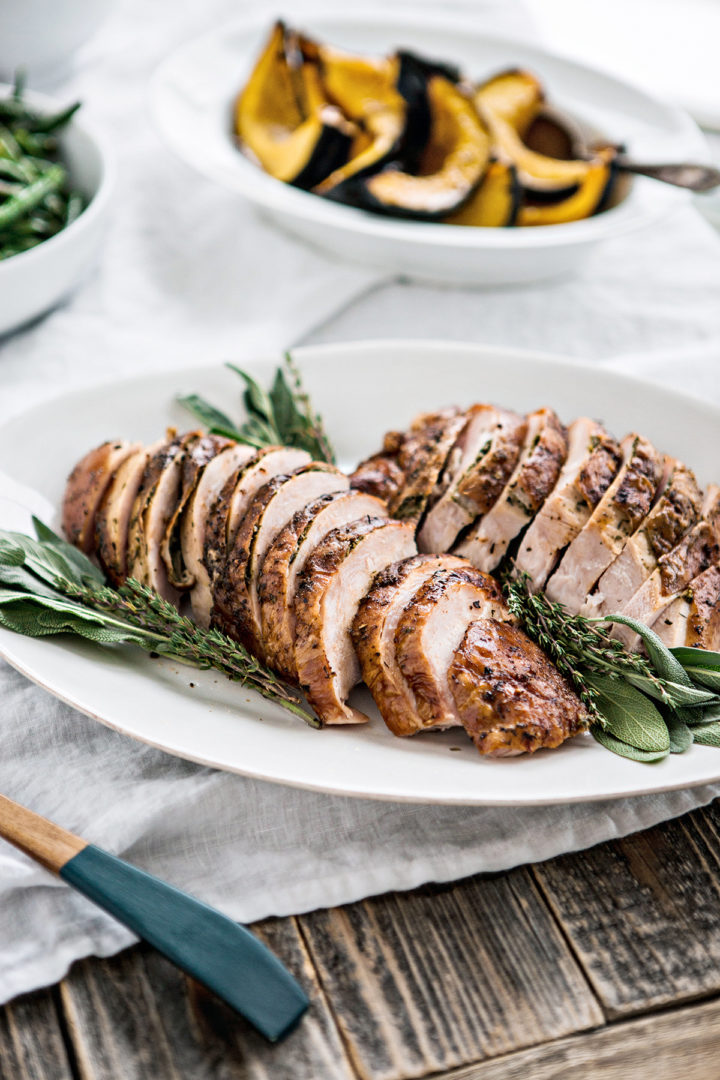 A white platter on a thanksgiving table with sliced smoked turkey fresh herbs. 