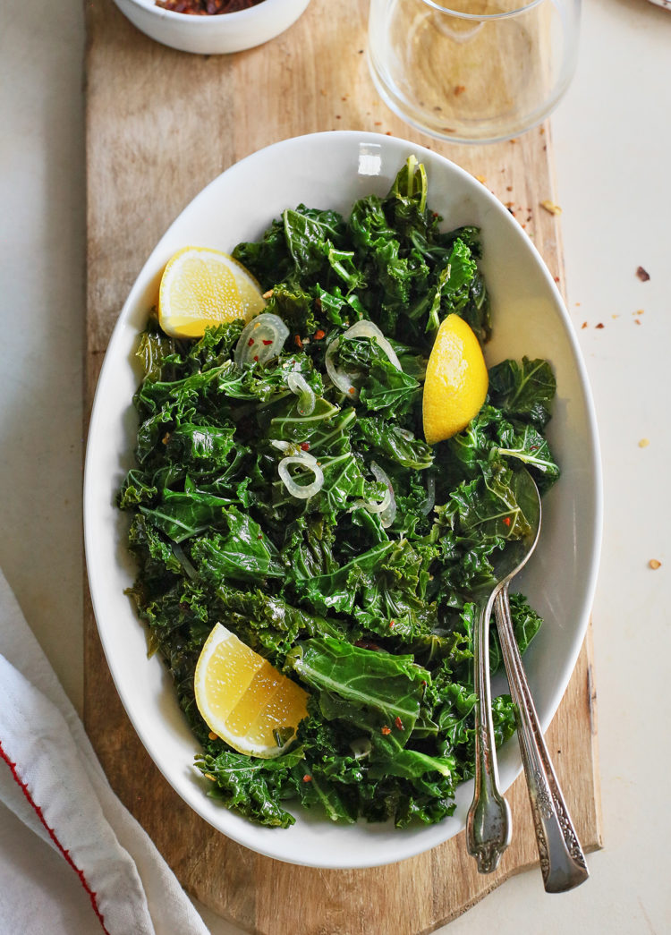 a white bowl of garlic sauteed kale on a wooden cutting board with a wine glass