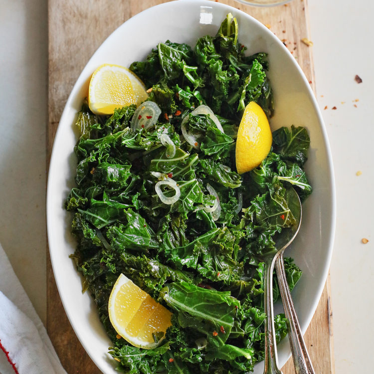 a white bowl of garlic sauteed kale on a wooden cutting board with a wine glass