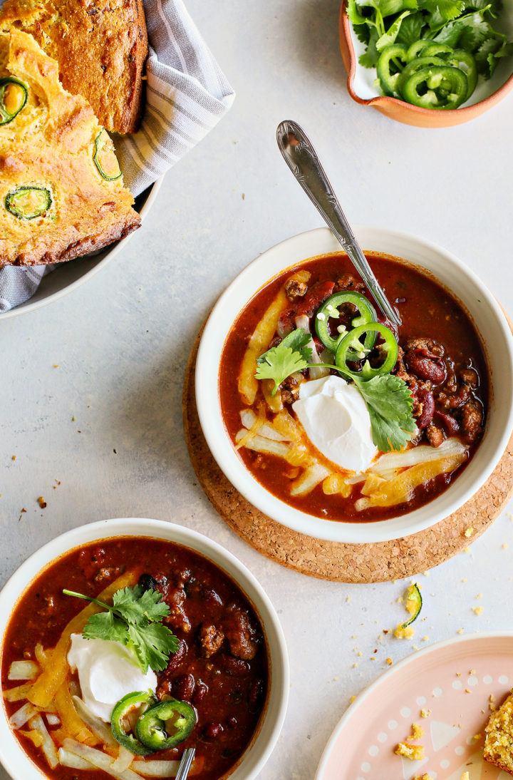 two bowls of ground beef chili with kidney beans next to a bowl of cornbread