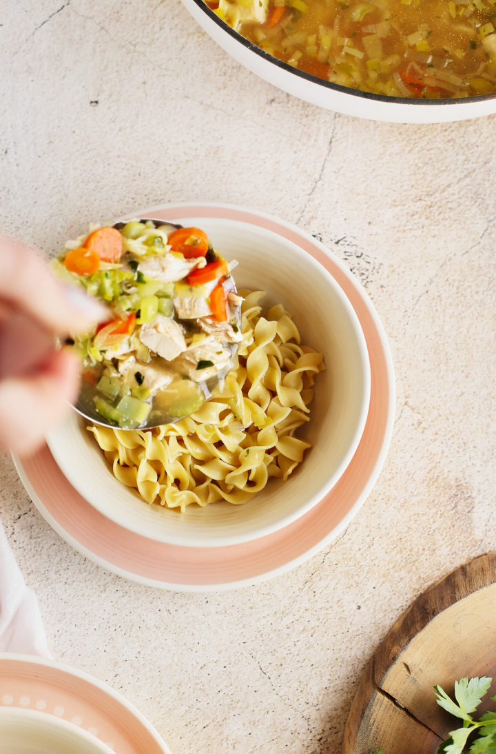 woman ladling homestyle chicken noodle soup into a bowl of egg noodles