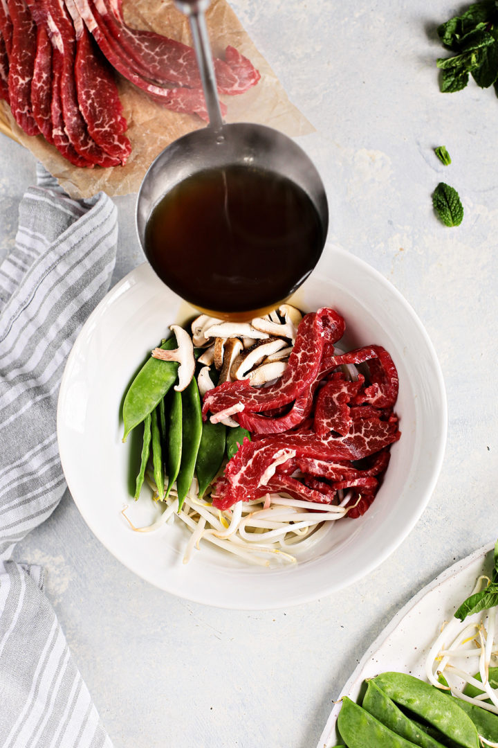 woman ladling hot broth over beef and rice noodles in a white bowl