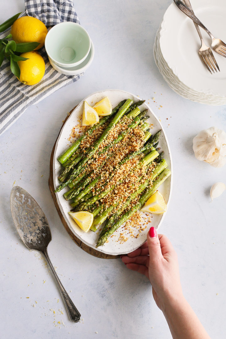 woman holding a white platter of the best grilled asparagus with parmesan panko breadcrumb topping and lemon wedges
