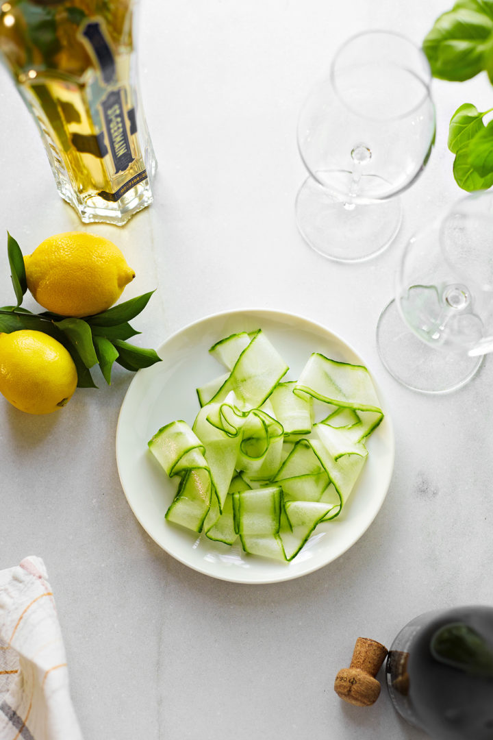 sliced cucumber ribbons and fresh lemons next to a bottle of st germain and wine glasses on a marble counter