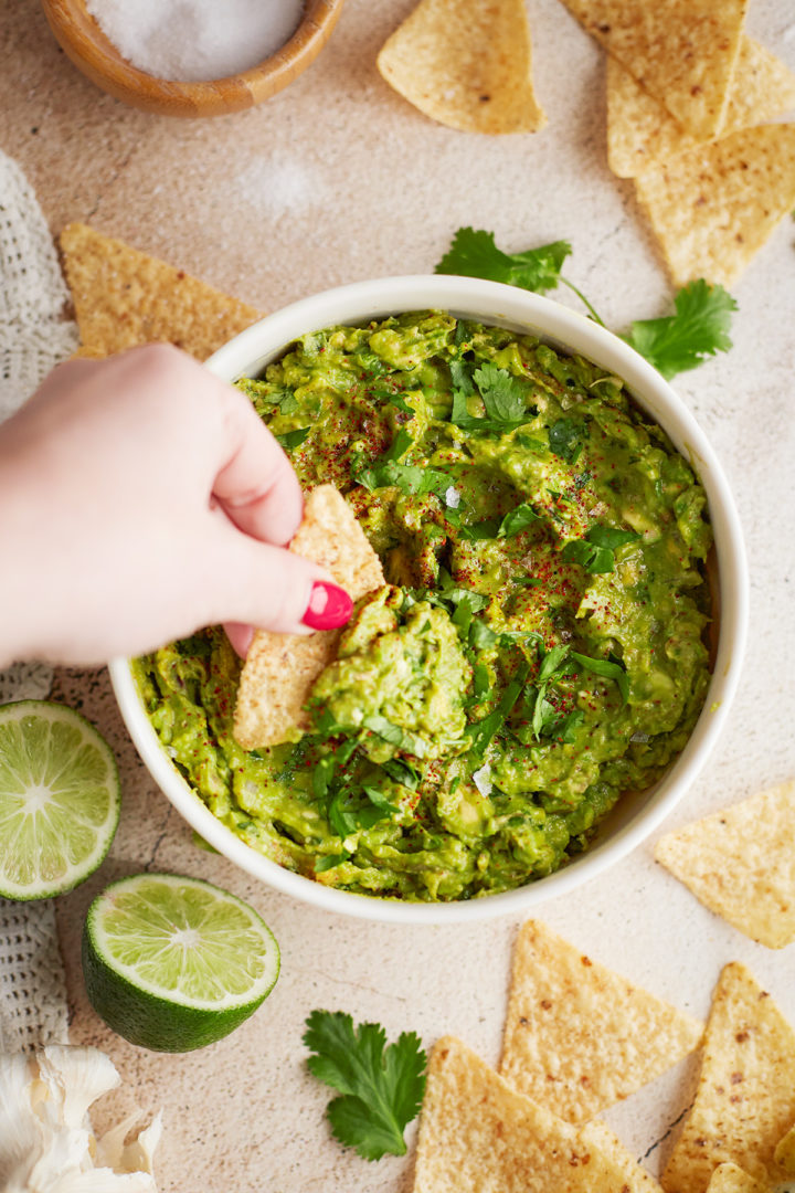 woman scooping homemade guacamole with a tortilla chip from a white serving bowl