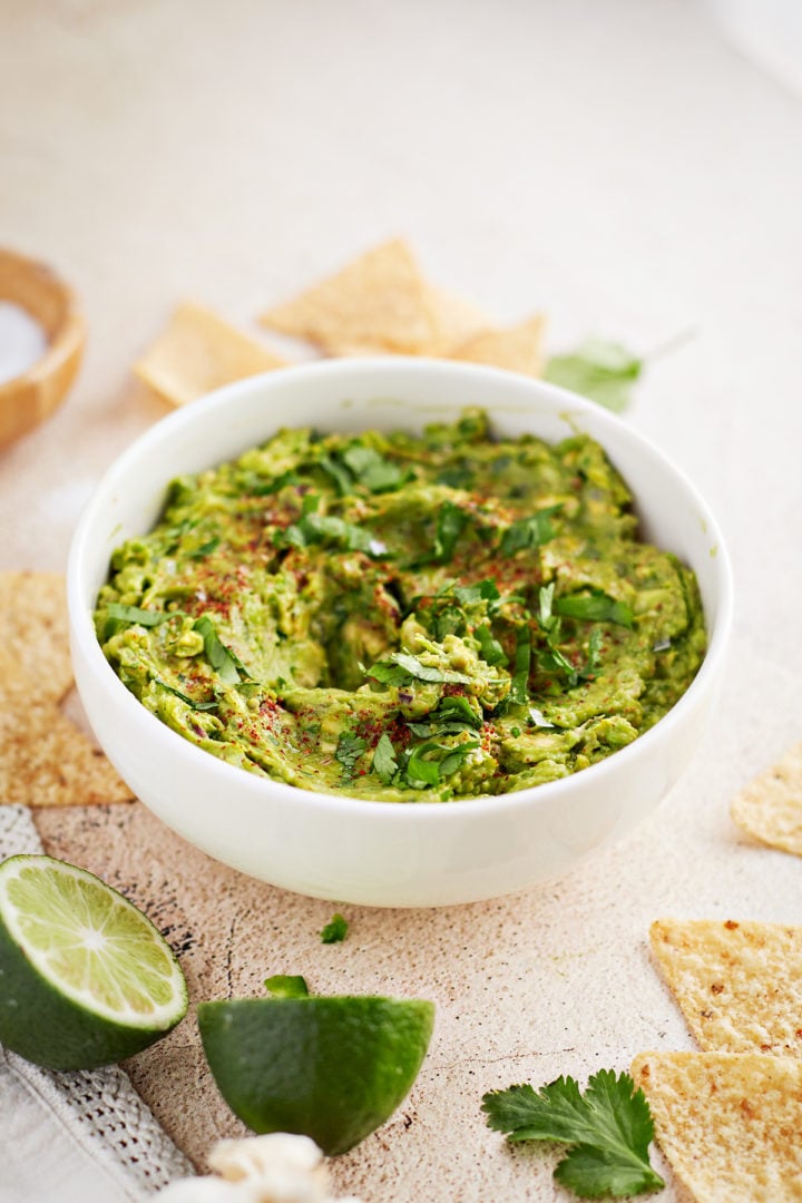 A white bowl filled with chunky guacamole garnished with chopped cilantro and a dusting of spices sits on a textured surface. Surrounding the bowl are tortilla chips, halved limes, fresh cilantro leaves, a garlic bulb, and a wooden salt bowl in the background. The guacamole has a rustic, hand-mixed texture and vibrant green color, signaling its freshness.