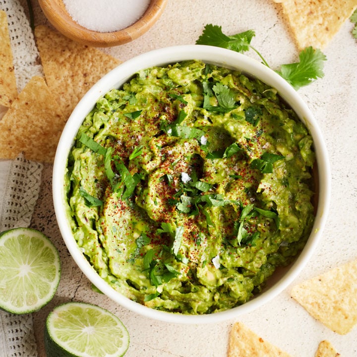 Finished guacamole overhead: Overhead shot of a bowl of simple guacamole garnished with fresh cilantro and chili powder. Surrounded by lime wedges, garlic, and tortilla chips.