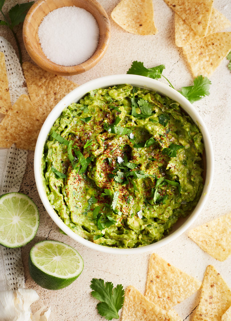 Finished guacamole overhead: Overhead shot of a bowl of simple guacamole garnished with fresh cilantro and chili powder. Surrounded by lime wedges, garlic, and tortilla chips.
