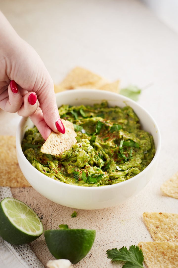 Side angle chip dip: A chip being dipped into a bowl of homemade guacamole, mid-scoop, with cilantro garnish and visible lime wedges in the background.