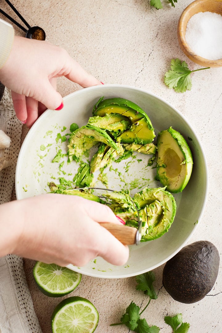 Mashing avocado in bowl: A ceramic bowl filled with partially mashed avocado being worked with a metal pastry blender. Fresh lime halves, cilantro, and garlic are scattered nearby.