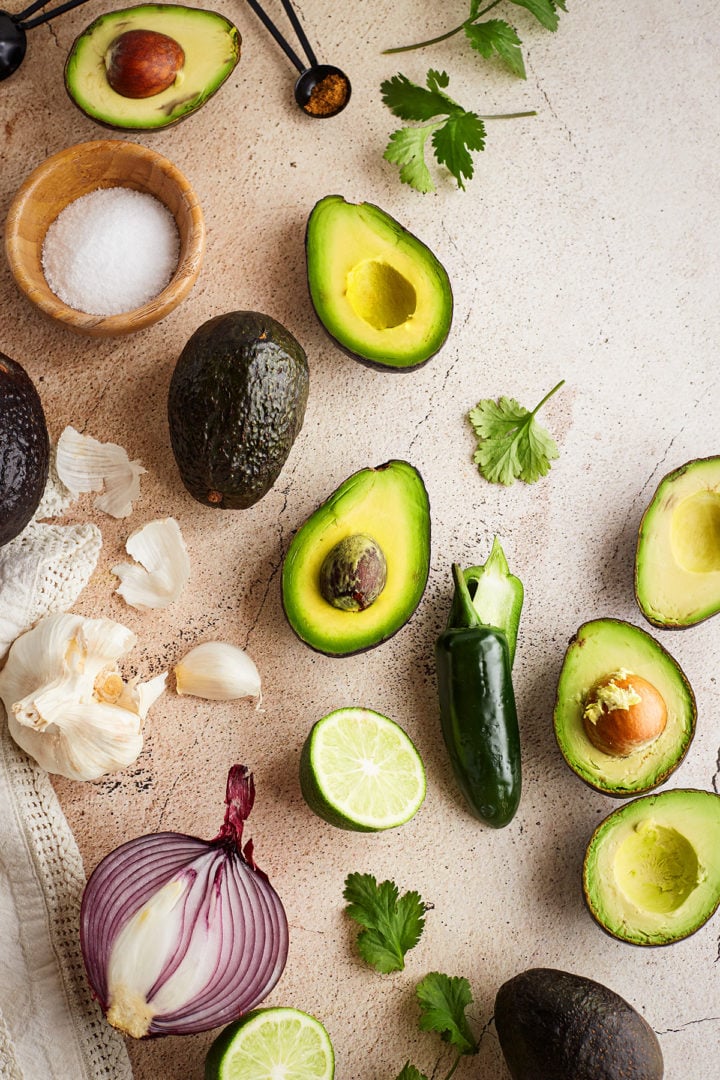 Guacamole ingredients flat lay: A visual spread of halved avocados, garlic cloves, jalapeño, halved limes, red onion, fresh cilantro, and spices laid out on a tan stone surface.
