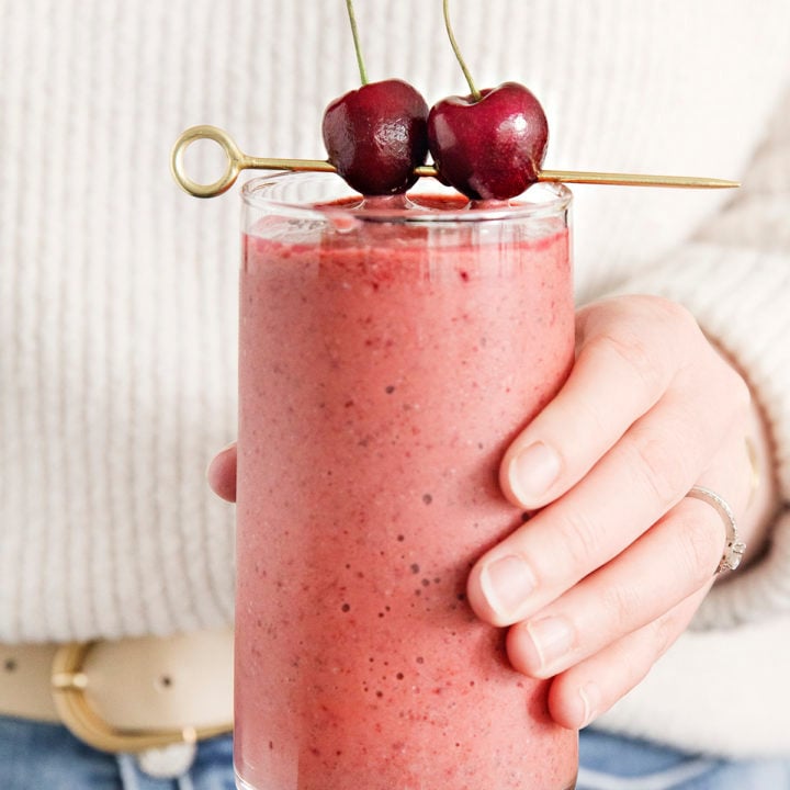 woman holding a glass of mango cherry smoothie garnished with fresh cherries on a cocktail skewer