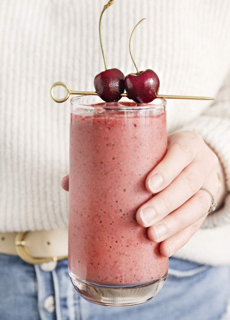 woman holding a glass of mango cherry smoothie garnished with fresh cherries on a cocktail skewer