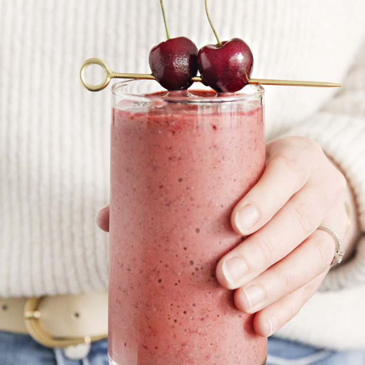 woman holding a glass of mango cherry smoothie garnished with fresh cherries on a cocktail skewer