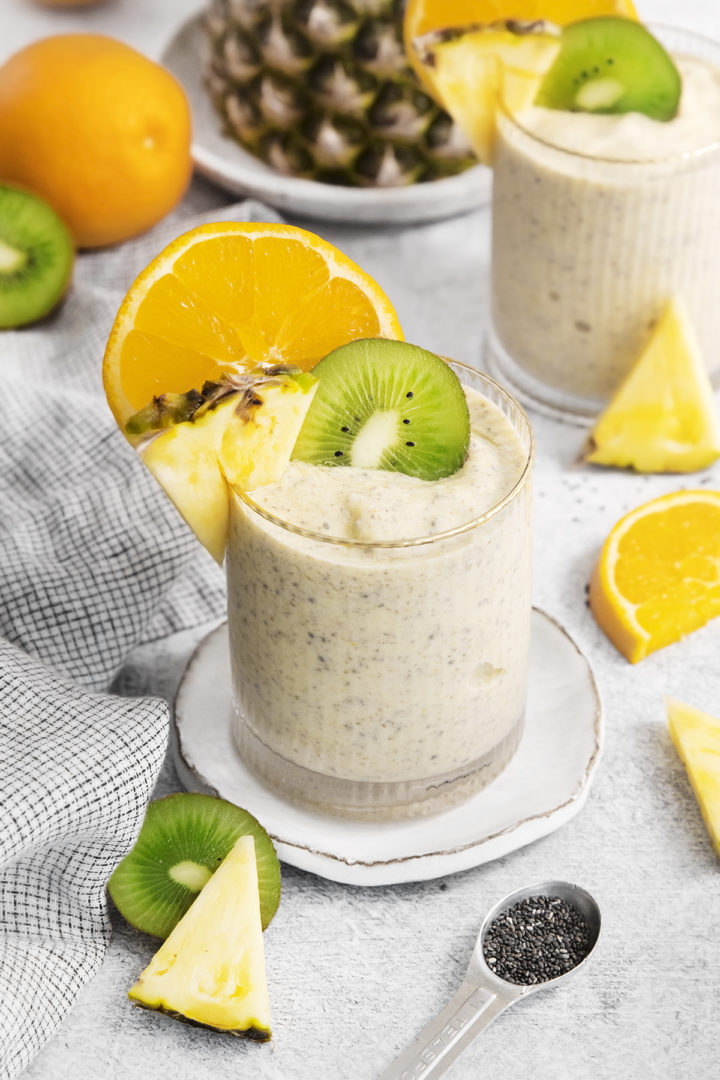 pineapple orange smoothie in two glasses surrounded by fresh pineapple, oranges, and kiwi