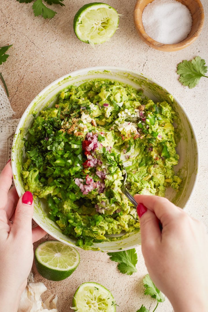 Stirring in ingredients: A spoon stirs fresh chopped cilantro, red onion, jalapeño, garlic, and spices into mashed avocado. A juiced lime, salt bowl, and herbs surround the bowl.