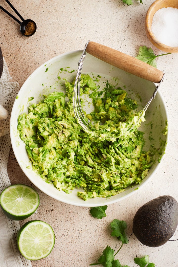 A white ceramic bowl filled with roughly mashed avocado sits on a light stone surface. A metal pastry blender with a wooden handle rests inside the bowl, partially covered in avocado. Around the bowl are halved limes, a whole avocado, fresh cilantro sprigs, and a pinch bowl of salt. A black measuring spoon with cumin powder is visible in the upper corner, suggesting the guacamole is mid-prep.