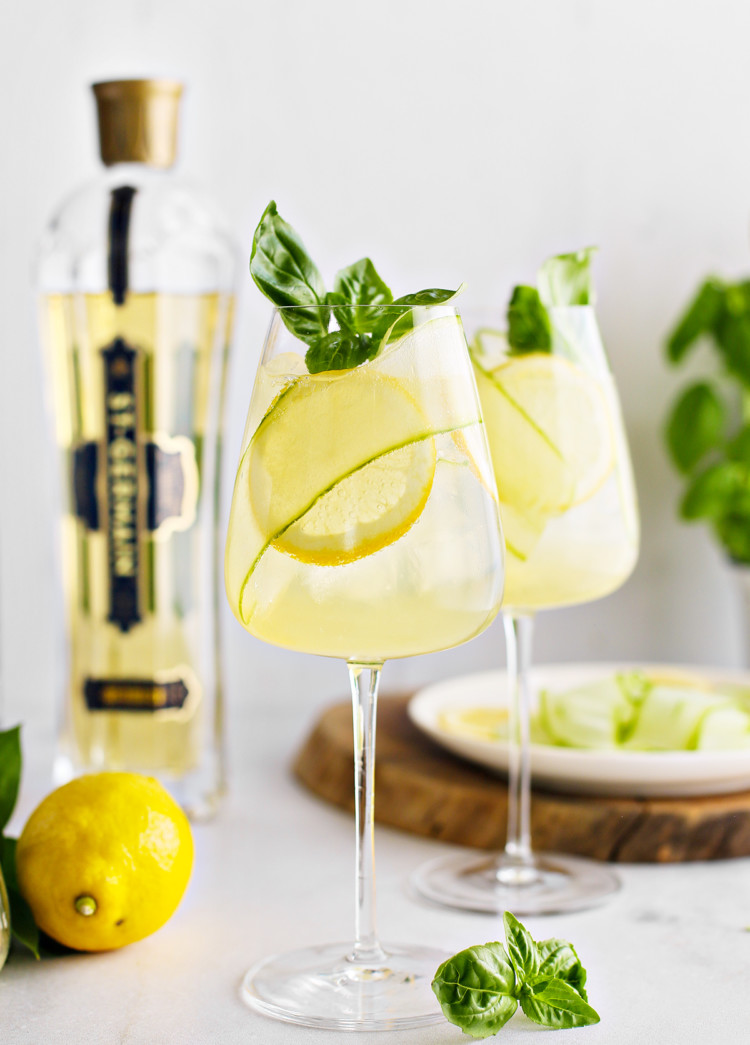 two glasses of st germain spritz garnished with lemon slices, cucumber ribbon, and fresh basil