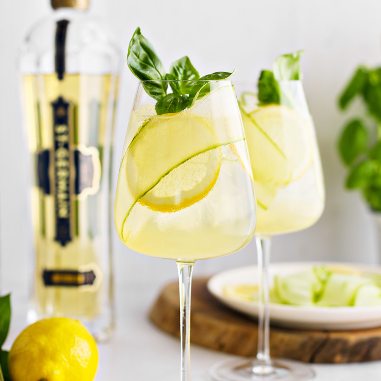two glasses of st germain spritz garnished with lemon slices, cucumber ribbon, and fresh basil