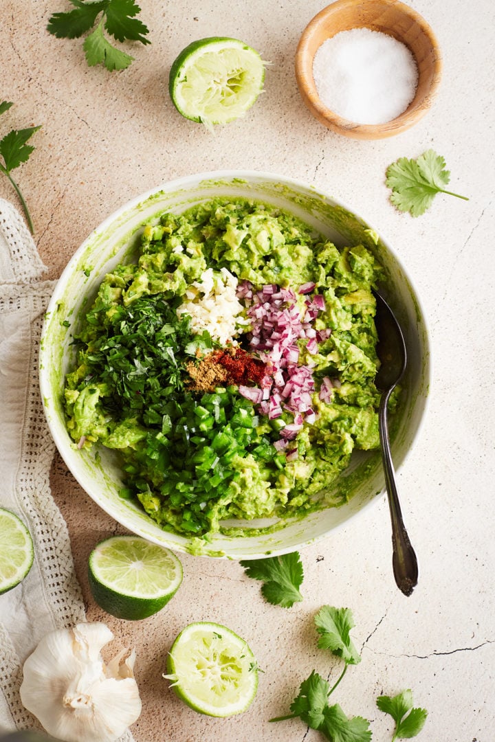 Stirring guacamole with full ingredients: A bowl of mashed avocado with separate piles of finely chopped garlic, onion, cilantro, jalapeño, and spices before mixing.