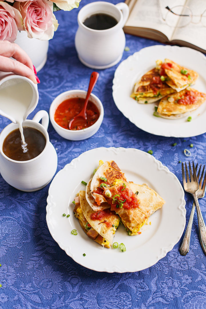 woman pouring cream into a mug of coffee next to a plate of breakfast quesadillas
