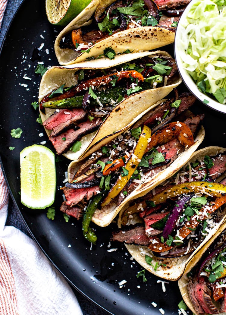 flank steak fajitas on a black plate with a bowl of lettuce and fresh limes