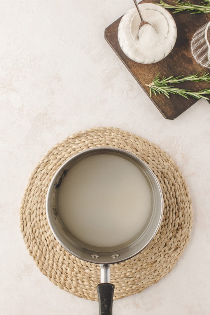 water and sugar in a stainless steel saucepan next to a bowl of sugar and fresh rosemary