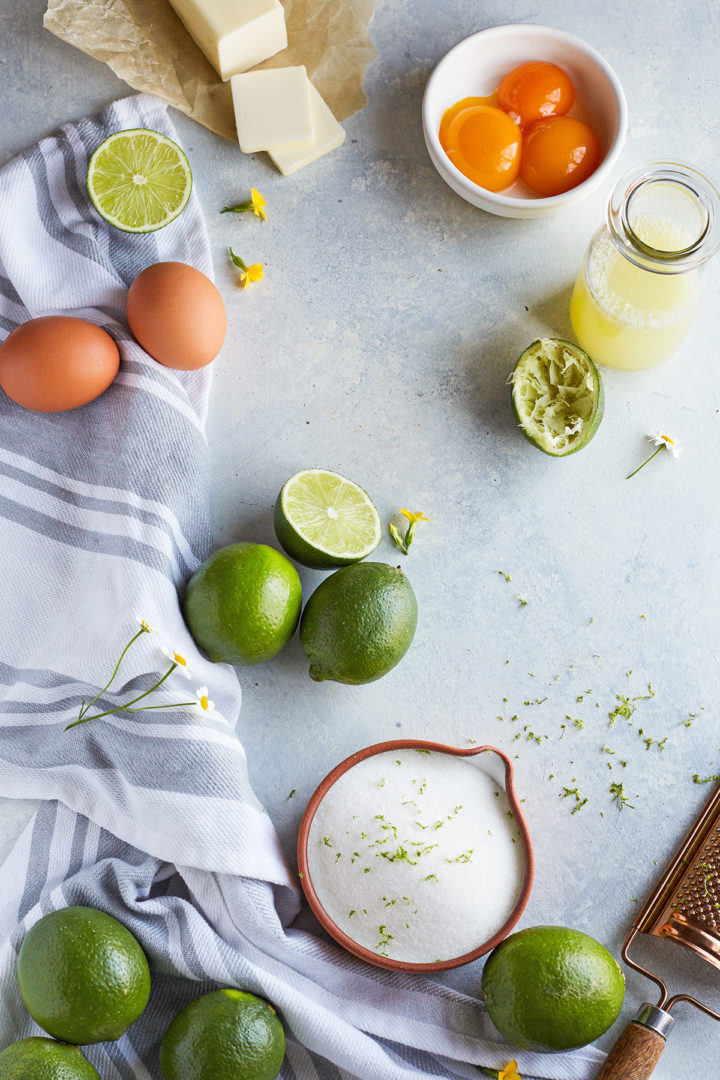 ingredients needed to make lime curd on a light blue background with a striped kitchen towel