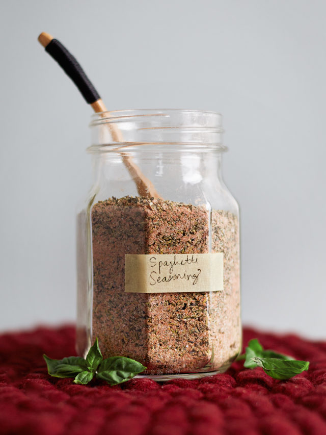 A glass jar filled with homemade spaghetti seasoning sits on a red felt trivet atop a rustic wooden table with a small wooden spoon resting inside, ready for scooping