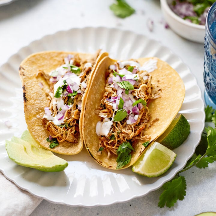 Two corn tortillas filled with tender Instant Pot shredded chicken taco meat, topped with chopped red onion, fresh cilantro, creamy cilantro lime crema, and avocado slices. The tacos are served on a scalloped white plate with lime wedges, surrounded by scattered herbs, sliced avocado, and blue drinking glasses. A bowl of red onion and cilantro, along with a plate of tortillas, can be seen in the softly blurred background—capturing a fresh, inviting setup for Instant Pot chicken tacos.