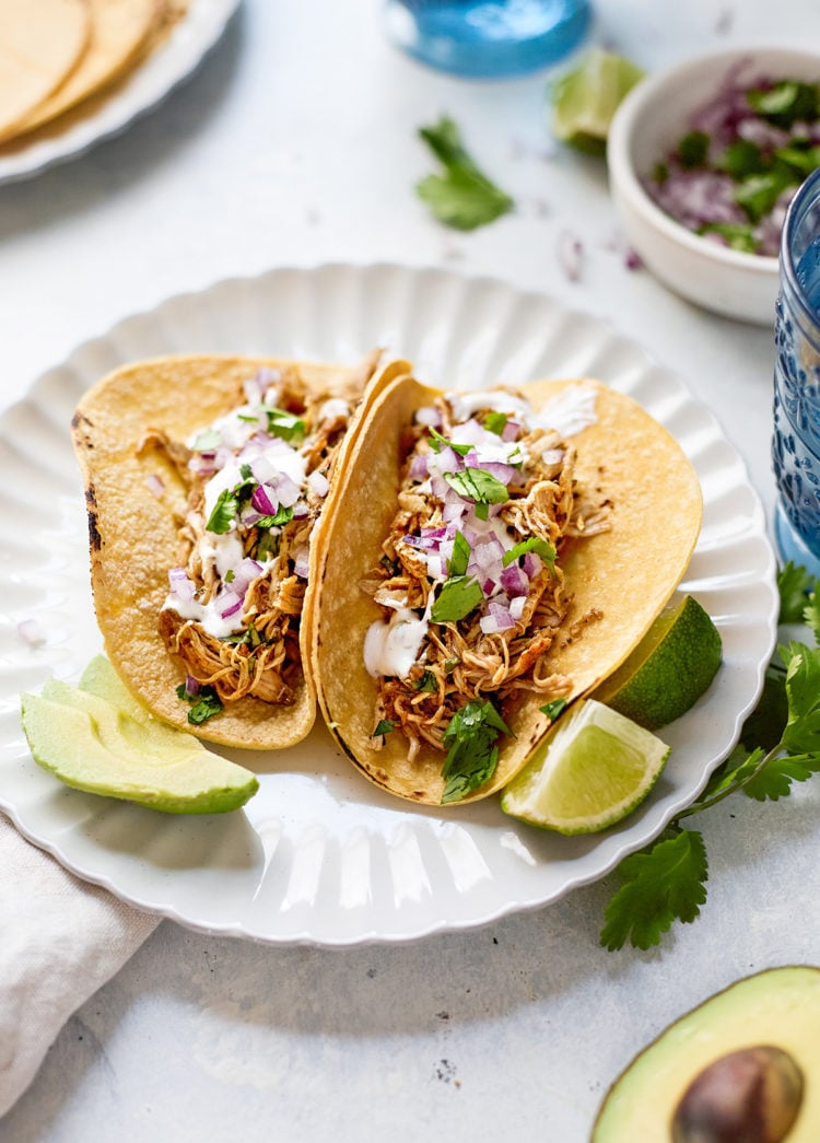Two corn tortillas filled with tender Instant Pot shredded chicken taco meat, topped with chopped red onion, fresh cilantro, creamy cilantro lime crema, and avocado slices. The tacos are served on a scalloped white plate with lime wedges, surrounded by scattered herbs, sliced avocado, and blue drinking glasses. A bowl of red onion and cilantro, along with a plate of tortillas, can be seen in the softly blurred background—capturing a fresh, inviting setup for Instant Pot chicken tacos.
