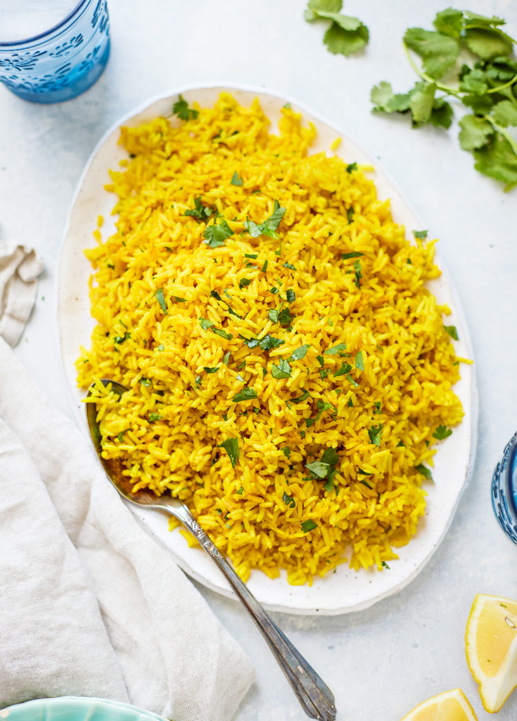Turmeric yellow rice on an oval white platter, with fresh cilantro and a kitchen towel nearby. S spoon rests on the plate, ready to serve.