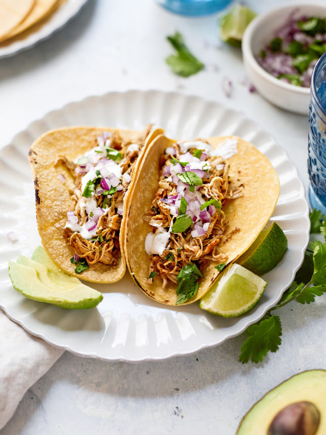 Two corn tortillas filled with tender Instant Pot shredded chicken taco meat, topped with chopped red onion, fresh cilantro, creamy cilantro lime crema, and avocado slices. The tacos are served on a scalloped white plate with lime wedges, surrounded by scattered herbs, sliced avocado, and blue drinking glasses. A bowl of red onion and cilantro, along with a plate of tortillas, can be seen in the softly blurred background—capturing a fresh, inviting setup for Instant Pot chicken tacos.