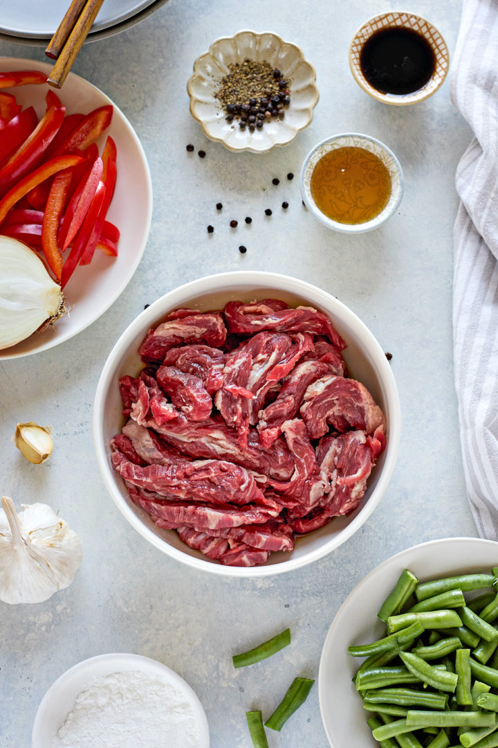 Overhead shot of black pepper beef ingredients arranged on a light blue surface, including thinly sliced raw beef, bright green beans, sliced red bell pepper strips, sliced white onions, minced garlic, minced ginger, and small bowls holding black pepper, dry sherry, and coconut aminos.