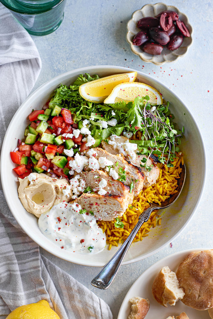 Overhead layout of meal-prepped chicken tzatziki bowls in glass containers, each with rice, grilled chicken, fresh veggies, tzatziki, and lemon slices for garnish.
