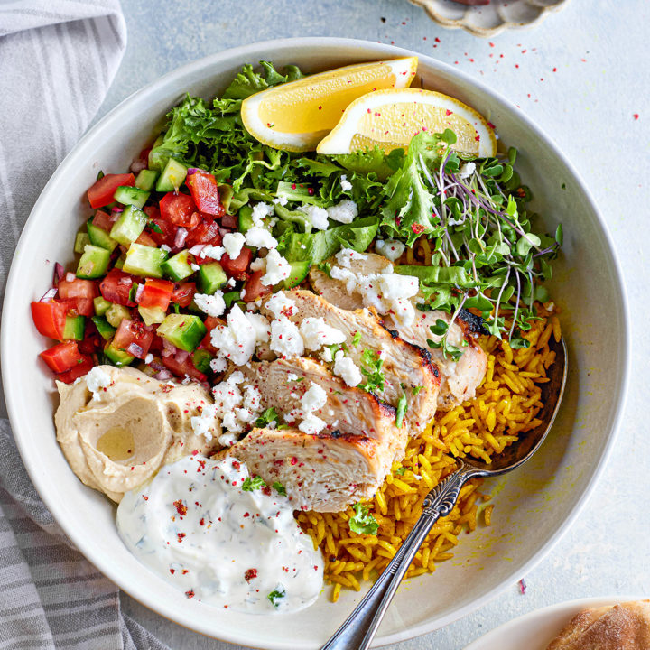 Overhead view of a white bowl filled with turmeric rice, grilled chicken slices, diced cucumber and tomatoes, red onion, tzatziki, hummus, and crumbled feta. A lemon wedge garnishes the bowl. A small bowl of olives and a linen napkin sit nearby.