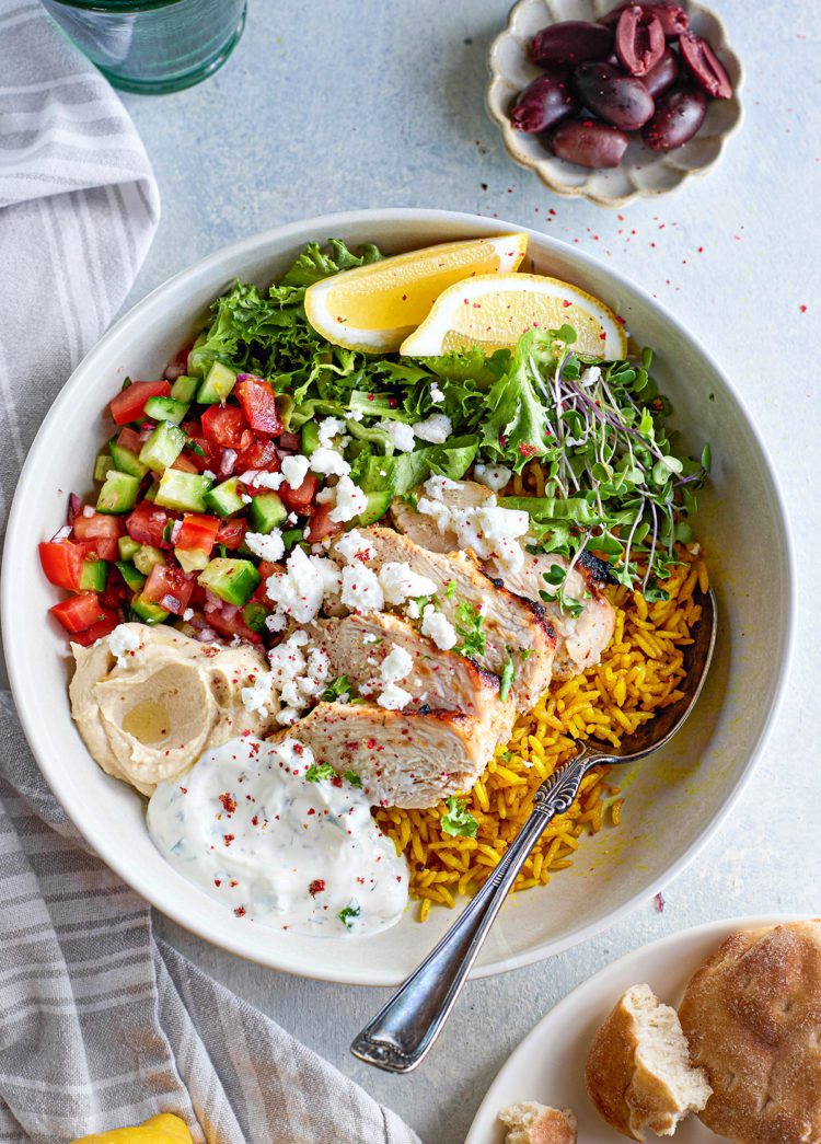 Overhead view of a white bowl filled with turmeric rice, grilled chicken slices, diced cucumber and tomatoes, red onion, tzatziki, hummus, and crumbled feta. A lemon wedge garnishes the bowl. A small bowl of olives and a linen napkin sit nearby.