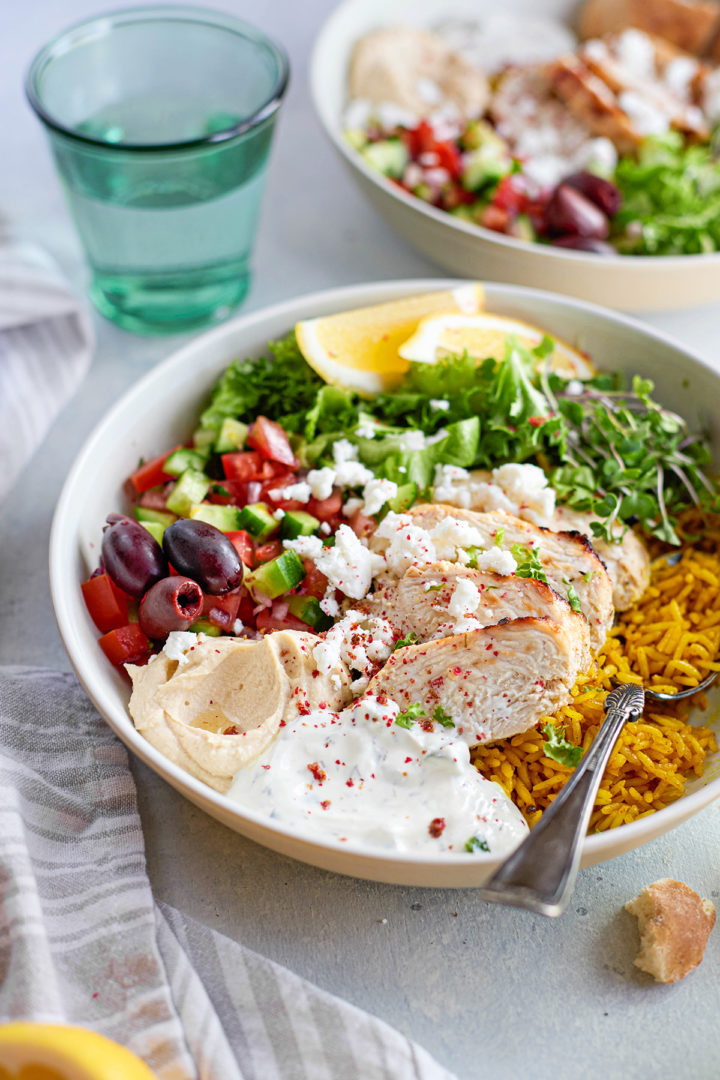Overhead image showing two chicken tzatziki rice bowls next to each other, each built with a base of turmeric rice and finished with colorful Greek-inspired toppings.
